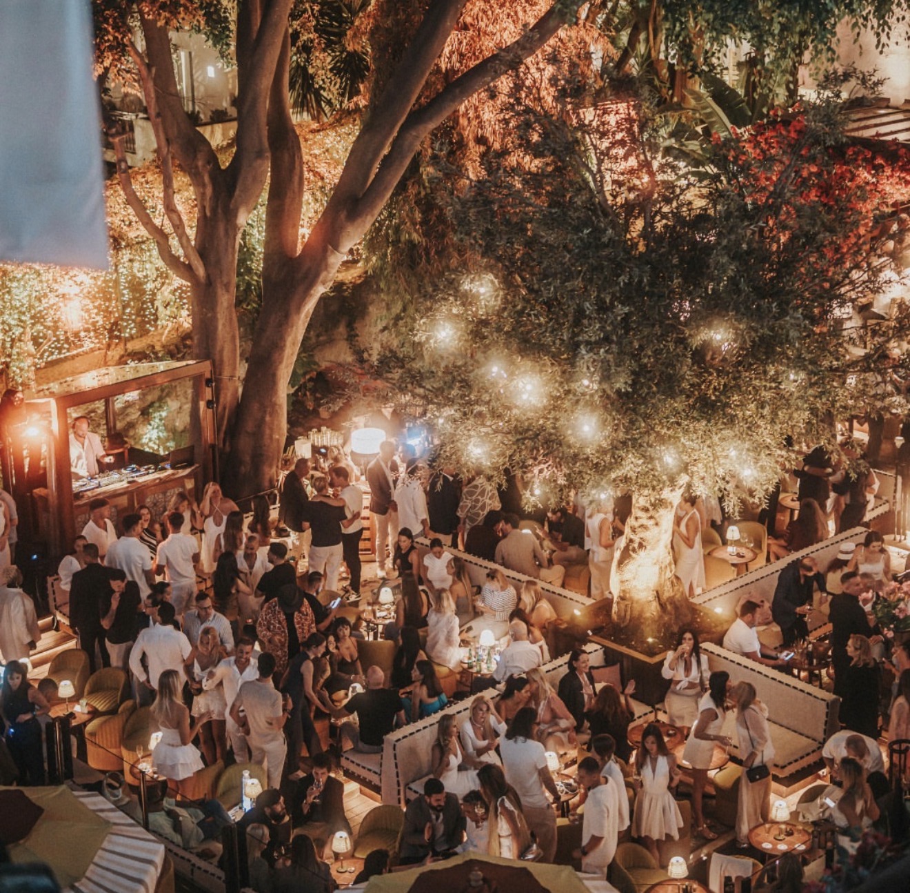 La Plaza at Puente Romano Marbella at night, crowd at lounge tables beneath lit fig trees